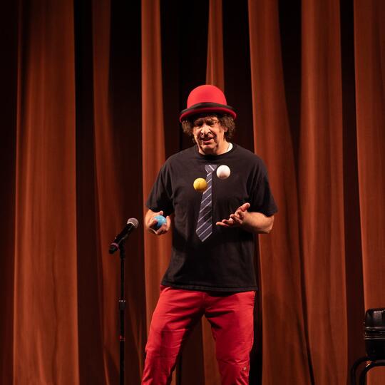 Marcus Wilson, in a red cap and jacket, juggles orange balls amid costumed entertainers, including stilt walkers, pirates, and a cowboy, at a lively event.